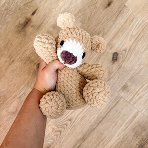 May include: A handmade, tan-colored crochet bear with a white face and a dark purple nose and mouth. The bear has small, round ears and is held in a hand. The bear's arms and legs are also round and plush.