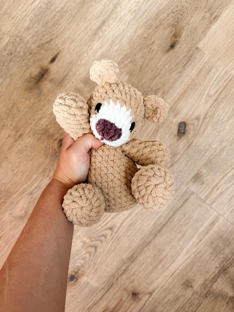 May include: A handmade, tan-colored crochet bear with a white face and a dark purple nose and mouth. The bear has small, round ears and is held in a hand. The bear's arms and legs are also round and plush.
