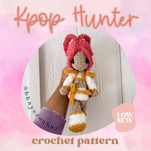 May include: A handmade crochet doll with a pink hairstyle, wearing a white and gold outfit. The doll has black eyes and is held in front of a pink background. Text on the image reads "Kpop Hunter" and "crochet pattern".