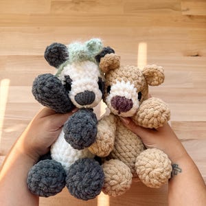 May include: Two handmade crochet bear plushies. One bear is white, gray, and black with a mint green tuft of hair. The other bear is tan with a purple nose. Both have round, fluffy bodies and are held up by hands.