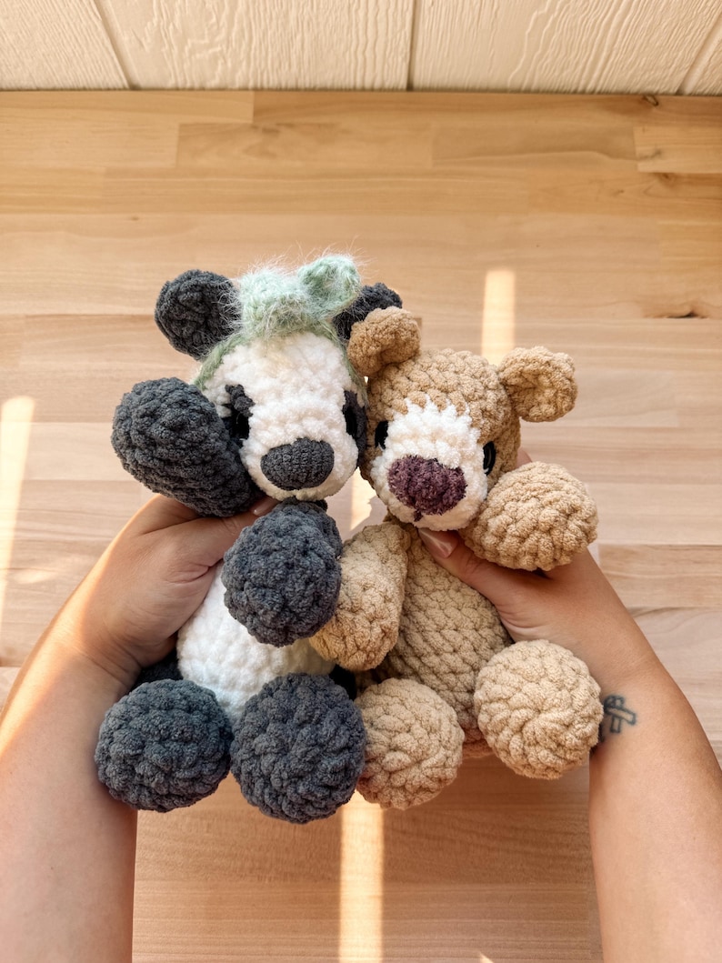May include: Two handmade crochet bear plushies. One bear is white, gray, and black with a mint green tuft of hair. The other bear is tan with a purple nose. Both have round, fluffy bodies and are held up by hands.