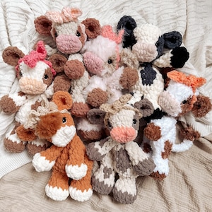 May include: A group of crocheted cow plushies in various colors and patterns, including brown, white, black, and pink. The plushies are arranged in a circle on a white knitted blanket.