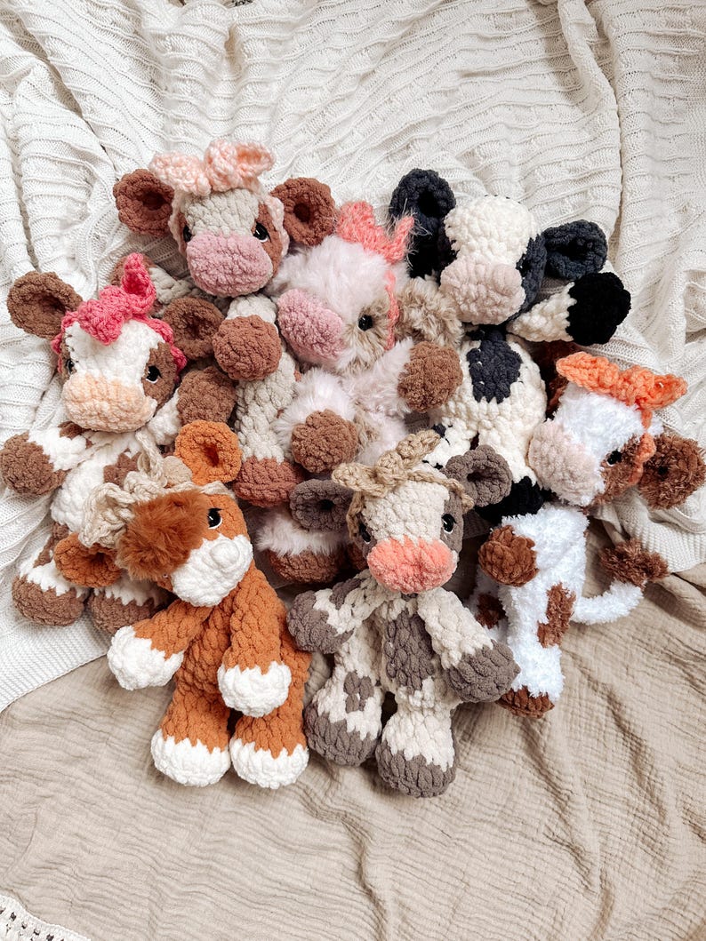 May include: A group of crocheted cow plushies in various colors and patterns, including brown, white, black, and pink. The plushies are arranged in a circle on a white knitted blanket.