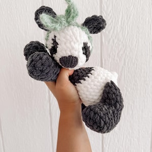 May include: A handmade crochet cow plushie in white, gray, and green. The cow has a white body with gray spots, gray hooves, and a gray and white face. The ears and the top of the head are green. The plushie is soft and cuddly.