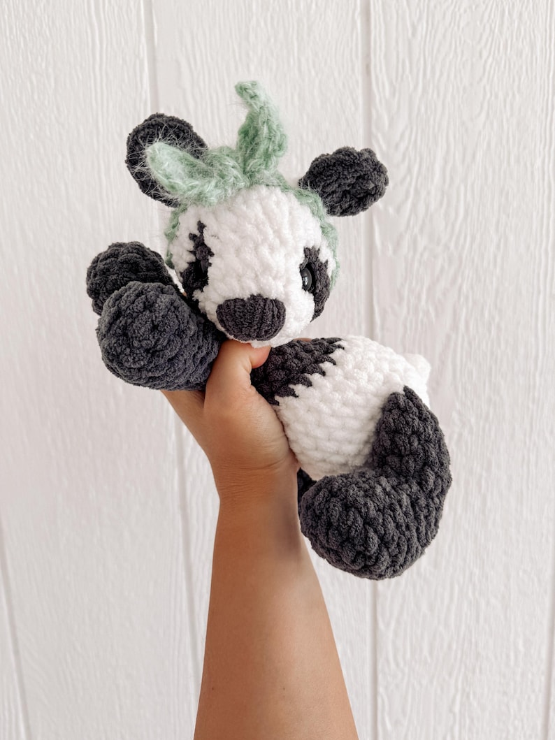 May include: A handmade crochet cow plushie in white, gray, and green. The cow has a white body with gray spots, gray hooves, and a gray and white face. The ears and the top of the head are green. The plushie is soft and cuddly.