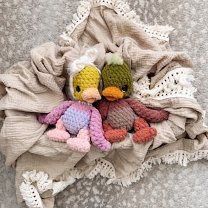 May include: Two handmade crochet duck toys with colorful bodies and orange beaks rest on a beige blanket with fringe. One duck has a yellow head with a white bow, the other has a green head with a green pom-pom.