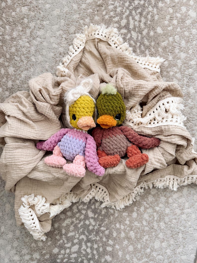 May include: Two handmade crochet duck toys with colorful bodies and orange beaks rest on a beige blanket with fringe. One duck has a yellow head with a white bow, the other has a green head with a green pom-pom.