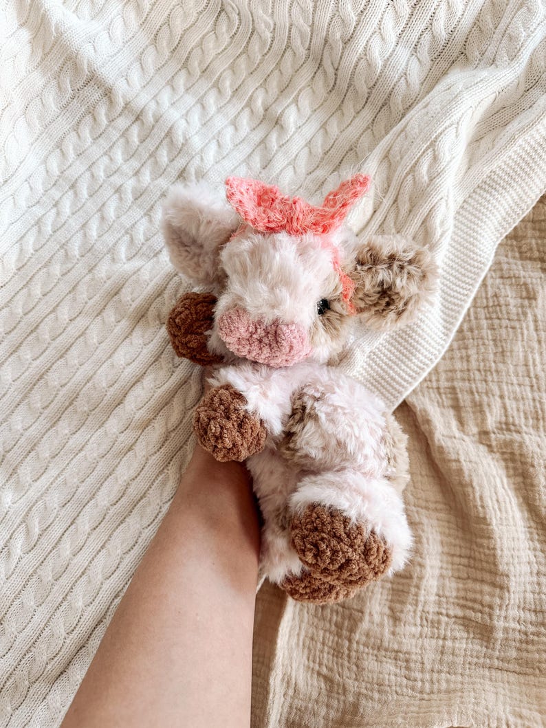 May include: A crocheted plush toy cow with brown and white fur, a pink nose, and a pink bow. The cow is lying on a white knitted blanket.