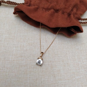 May include: A gold chain necklace with a clear round pendant. The necklace is lying on a beige fabric surface.