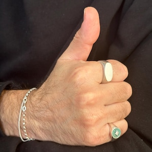 May include: A silver bracelet and two silver rings are visible on a hand giving a thumbs up. One ring has an oval face, and the other has a green face with a star design.