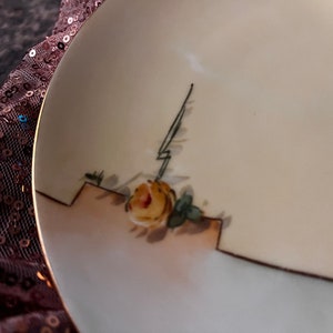 May include: A white ceramic plate with a gold rim and a hand-painted yellow rose with green leaves. The rose is in the lower right corner of the plate.