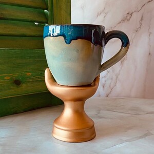 May include: A ceramic mug with a blue and white glaze sits on a gold stand. The mug has a rounded shape and a handle.