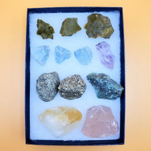 May include: A collection of raw gemstones in a black box with white padding. The gemstones include green, blue, purple, gold, and pink stones. The box is rectangular and sits on a light orange surface.