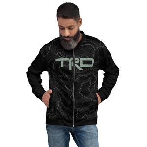 May include: Black bomber jacket with a topographic map design and the letters "TRD" in a faded green color. The jacket has a zipper closure and two front pockets. The jacket is worn by a person.