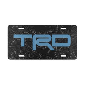May include: Rectangular license plate with a black background featuring a white topographic map design. The letters "TRD" are in a light blue color. The plate has four mounting holes.