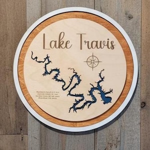 May include: A round wooden sign featuring a map of Lake Travis. The lake's outline is in blue, set against a light wood background, with the words "Lake Travis" in a script font. A compass rose is also included.