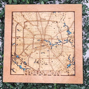 May include: A wooden map of Houston, Texas, with a light brown frame. The map features intricate laser-cut details of streets and waterways, with blue accents representing bodies of water. The city name "HOUSTON" is spelled out at the bottom.