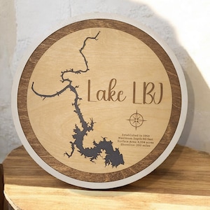 May include: Round wooden sign featuring a map of Lake LBJ, with the lake's outline in dark brown. The sign includes the text "Lake LBJ" and a compass rose. Additional text provides details about the lake's establishment, depth, area, and shoreline.