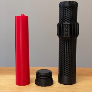 Puede incluir: Un conjunto de objetos impresos en 3D: un cilindro rojo brillante, una tapa negra roscada y un agarre cilíndrico negro texturizado. El agarre presenta un patrón de panal y una banda central con tres detalles circulares. Los objetos están sobre una superficie de madera.
