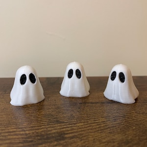 Op de afbeelding: Drie witte spookfiguren met zwarte ogen. De spoken staan op een houten oppervlak.
