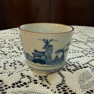 May include: A white ceramic cup with a blue landscape design. The cup has a slightly textured surface and a brown rim. The landscape features trees, buildings, and mountains. The cup is sitting on a white lace tablecloth.