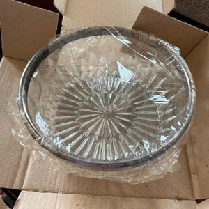 May include: A clear glass bowl with a silver rim, packaged in plastic wrap and placed inside a cardboard box. The bowl has a starburst pattern radiating from the center.