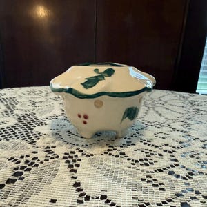 May include: A small, off-white ceramic container with a scalloped rim. The container is decorated with green leaf and bow designs, and small red dots. It has three small feet and is sitting on a white lace tablecloth.