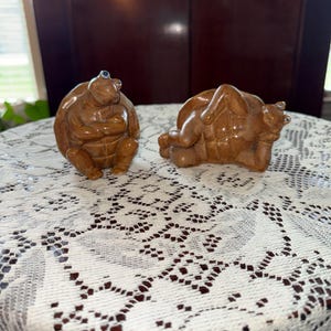 Puede incluir: Dos figuras de tortugas marrones. Una tortuga está sentada con los brazos cruzados, la otra está tumbada. Ambas tienen un caparazón texturizado y pequeños ojos azules. Las figuras están sobre un mantel de encaje blanco.