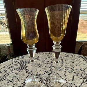 May include: Two elegant, vintage-style champagne flutes. The glasses have a clear, slender stem and a flared, amber-colored bowl. The flutes are displayed on a white lace tablecloth, suggesting a formal setting.