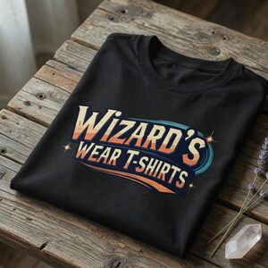 Pode incluir: Uma t-shirt preta de manga curta com um design gráfico que apresenta o texto "WIZARD'S WEAR T-SHIRTS" numa fonte estilizada de inspiração retro. As letras incorporam um gradiente de cores laranja, amarelo e azul com pequenas estrelas.