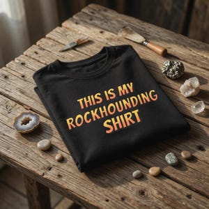 以下が含まれることがあります： オレンジと黄色の「THIS IS MY ROCKHOUNDING SHIRT」という文字が入った黒いTシャツ。木製の表面に折りたたまれており、周りには岩や道具が散らばっています。クルーネックのTシャツです。