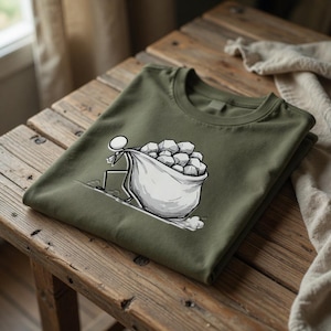 以下が含まれることがあります： オリーブグリーンのTシャツ。白黒の漫画のグラフィックで、棒人間が大きな石の袋を運んでいる様子が描かれています。Tシャツは木製の表面にきれいに折りたたまれています。
