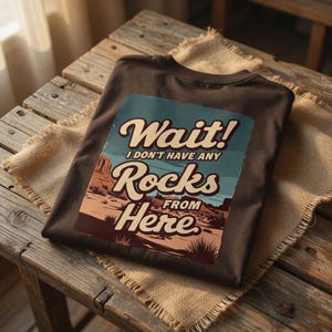 May include: A dark brown t-shirt with a graphic that reads "Wait! I don't have any Rocks From Here." The graphic features a desert landscape in shades of brown, tan, and blue. The shirt is folded on a wooden surface.