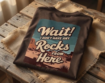 May include: A dark brown t-shirt with a graphic that reads "Wait! I don't have any Rocks From Here." The graphic features a desert landscape in shades of brown, tan, and blue. The shirt is folded on a wooden surface.