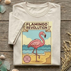 May include: A beige short-sleeved t-shirt featuring a graphic print of a pink flamingo standing on a beach. The text reads FLAMINGO REVOLUTION at the top and PROTECT THE FLAMINGO inside a circular emblem at the bottom of the rectangular design.