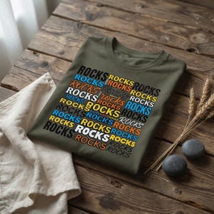 以下が含まれることがあります： 「ROCKS」の文字が格子状に繰り返されたオリーブグリーンのTシャツ。 「ROCKS」の文字は、青、オレンジ、白など様々な色で表示されています。 Tシャツは、麦の穂と石が置かれた木製の表面に折りたたまれています。