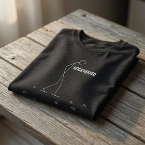 Pode incluir: Camiseta cinza escuro com um gráfico branco de um boneco de palito e a palavra "ROCKHOUND". O boneco de palito parece estar escalando ou trabalhando com pedras. A camiseta está dobrada sobre uma superfície de madeira.