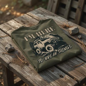 Könnte beinhalten: Ein olivgrünes T-Shirt mit dem Text "I'M HERE TO BREAK THINGS" und einer Grafik eines Geländewagens. Das Shirt liegt gefaltet auf einer verwitterten Holzoberfläche mit kleinen Steinen und Kiefernnadeln.