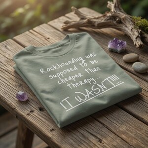 以下が含まれることがあります： 「Rockhounding was supposed to be cheaper than therapy... IT WASN'T!!!」の白文字がプリントされた、セージグリーンのTシャツ。木製の表面に折りたたまれ、アメジストの結晶と石が近くに置かれています。