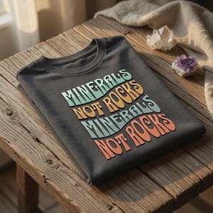 以下が含まれることがあります： レトロなフォントで「MINERALS NOT ROCKS」と書かれた濃い灰色のTシャツ。テキストは、ターコイズ、オレンジ、コーラルの色で重ねられています。木製の表面に折りたたまれたシャツで、背景にはクリスタルがあります。