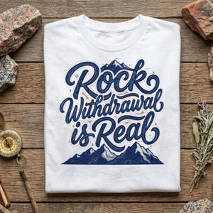 以下が含まれることがあります： 「Rock Withdrawal is Real」という文字がスタイリッシュな筆記体で描かれた、ダークブルーのグラフィックデザインが特徴の白いTシャツ。デザインには、地形の等高線が描かれた山の頂上のイラストと、小さなナビゲーションシンボルが含まれています。