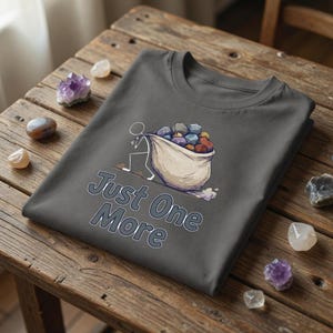 以下が含まれることがあります： 「Just One More」の文字と、カラフルな石が入った袋を運ぶ棒人間のイラストが描かれたダークグレーのTシャツ。木製の表面に折りたたまれており、周りにはクリスタルが散らばっています。