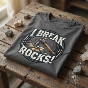 以下が含まれることがあります： 「I BREAK ROCKS!」の文字が円形デザインでプリントされたグレーのTシャツ。デザインは、岩を砕くツルハシが特徴です。Tシャツは、小さな石が散らばる木製の表面に折りたたまれています。