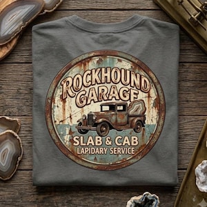 以下が含まれることがあります： チャコールグレーのTシャツで、大きな瑪瑙のスライスを積んだ錆びたヴィンテージトラックが描かれた円形のグラフィックが特徴です。テキストには「Rockhound Garage, Slab & Cab, Lapidary Service」と書かれています。デザインは風化したような、使い古された酸化した外観をしています。