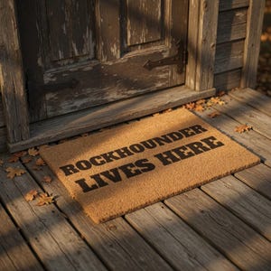 以下が含まれることがあります： 「ROCKHOUNDER LIVES HERE」の文字が黒でプリントされた長方形のコイア繊維製の玄関マット。風化した木製のドアの近く、木製のポーチに置かれています。