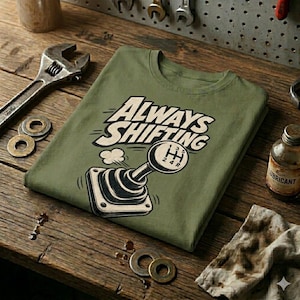 Könnte beinhalten: Ein olivgrünes T-Shirt mit dem Schriftzug "Always Shifting" und einer Grafik eines Autoschalthebels. Das T-Shirt liegt gefaltet auf einer Holzoberfläche, mit Werkzeugen und einer Schmierstoffflasche im Hintergrund.