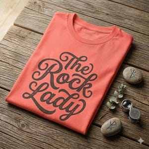 以下が含まれることがあります： コーラルカラーのクルーネックTシャツで、ダークグレーの筆記体風フォントで「The Rock Lady」という文字がプリントされています。シャツの横には、記号が刻まれた2つの滑らかなグレーの石と、小さな銀色の金属製ルーペが置かれています。