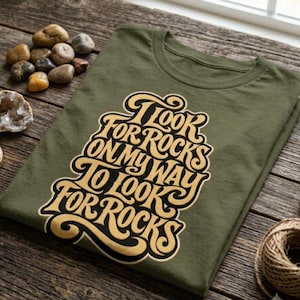 以下が含まれることがあります： オリーブグリーンのTシャツで、クリーム色と黒の大きなスタイリッシュなグラフィックプリントが施されています。テキストは「I LOOK FOR ROCKS ON MY WAY TO LOOK FOR ROCKS」と書かれており、衣服の中央に積み重ねられた渦巻くフォントデザインで配置されています。