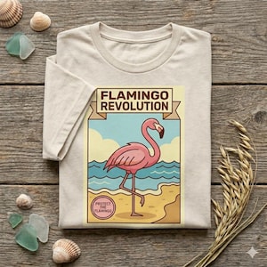 May include: A beige short-sleeve t-shirt featuring a graphic print of a pink flamingo standing on a beach. The text reads FLAMINGO REVOLUTION at the top and PROTECT THE FLAMINGO inside a circular badge at the bottom of the rectangular illustration.