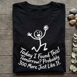 以下が含まれることがあります： 黒いクルーネックTシャツ。白い棒人間のグラフィックが小さな物体を持っているデザイン。その下に「Today I Found This! Tomorrow? Probably 300 More Just Like It.」という白いテキストがプリントされている。半袖の標準的なフィット感のTシャツ。